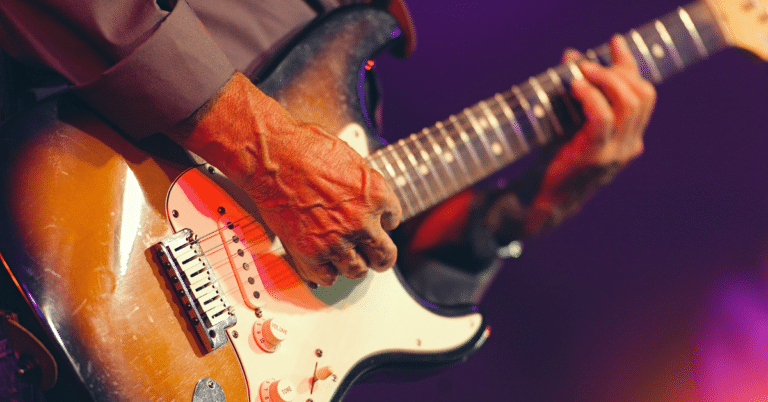 E-Gitarrist spielt auf großer Bühne eine Fender Strat – Fokus auf rechter Anschlagshand im Scheinwerferlicht. Headerbild für den E-Gitarrenkurs für Fortgeschrittene.