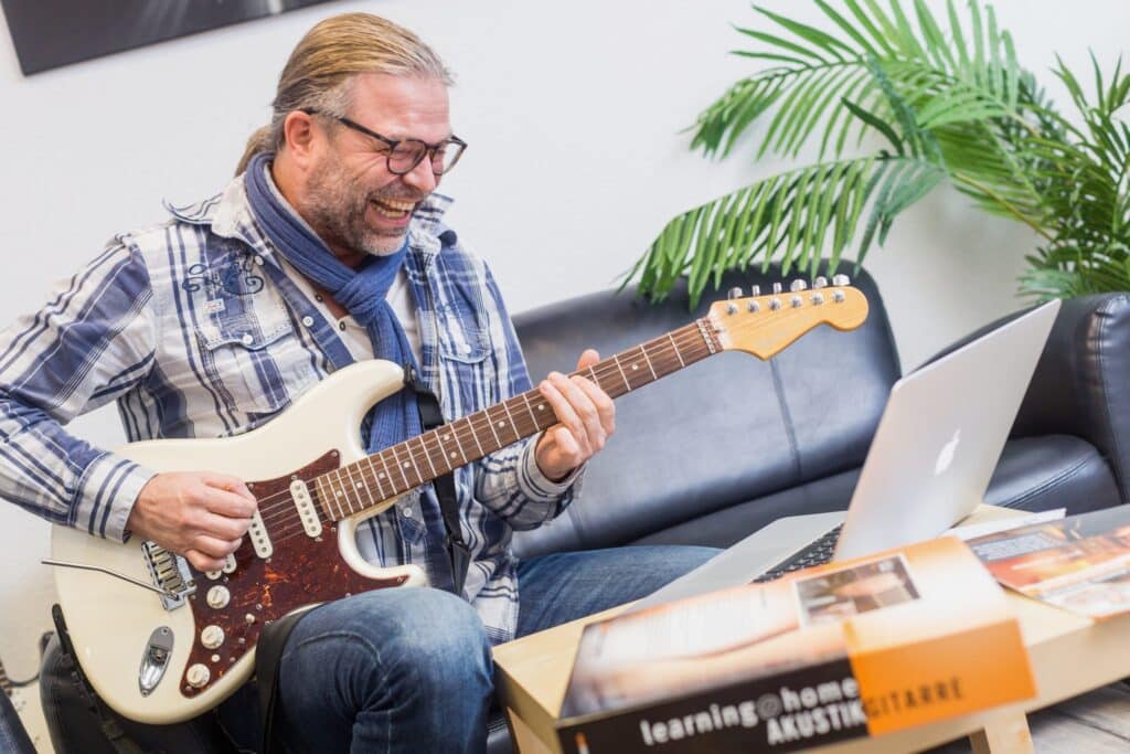 Musikstudium E-Gitarre. NMA-Professional-Student übt begeistert E-Gitarre auf der Couch vor dem Laptop – E-Gitarrenkurs für Profis mit Original-Unterlagenordner